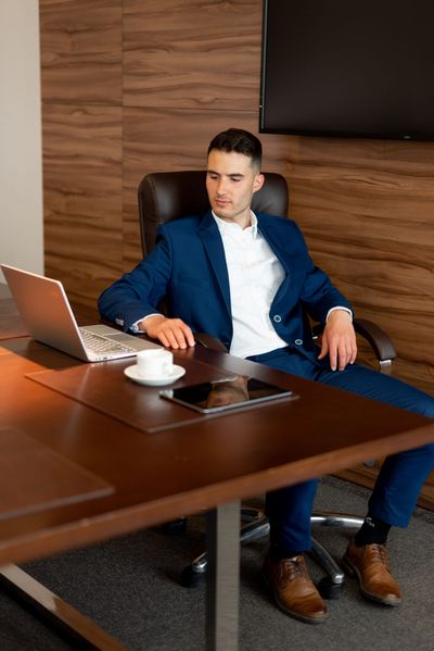 A business owner in a navy suit reclining at a boardroom desk, reviewing work on a laptop